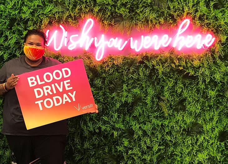 Blood Drive coordinator holding a sign next to a "Wish you were here" neon sign.